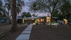 A modern house with a paved driveway, a covered patio, and a well-lit exterior at dusk. - Mid-century Modern W/backyard Oasis, Putting Green (Phoenix)
