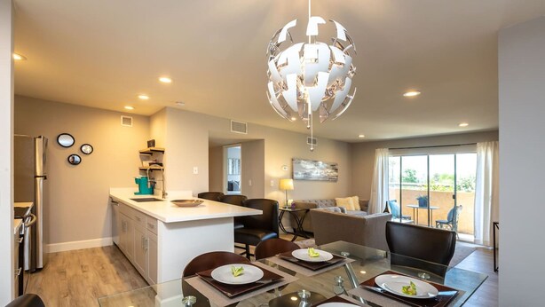Una cocina moderna con una isla central, comedor con mesa de vidrio y sala de estar con sofá y televisor.