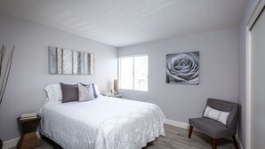 Un dormitorio con cama, una silla, una mesita de noche, una ventana y decoración en la pared.