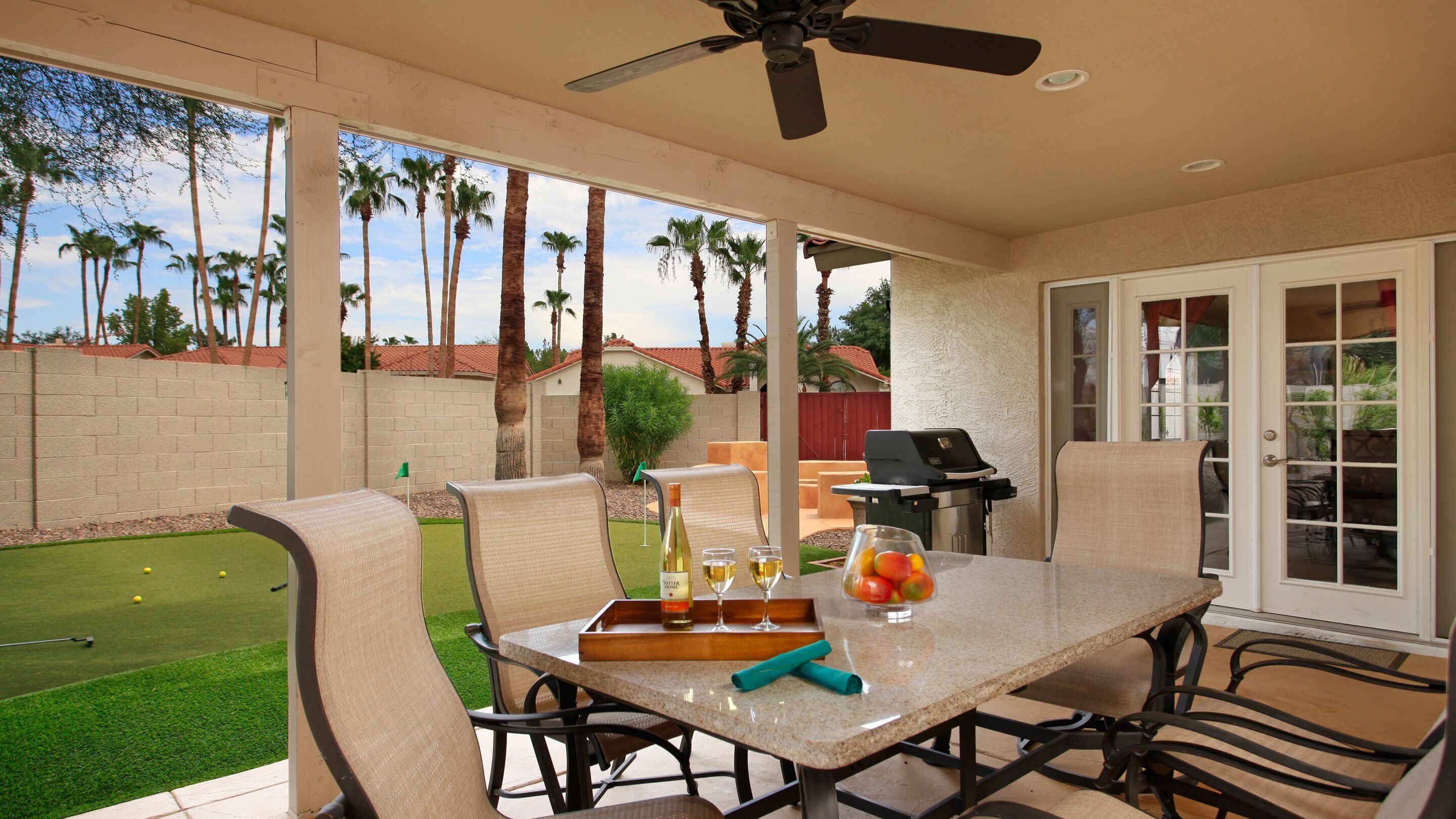 A patio area with a table set for dining, chairs, a ceiling fan, and a view of a backyard with a lawn and palm trees.