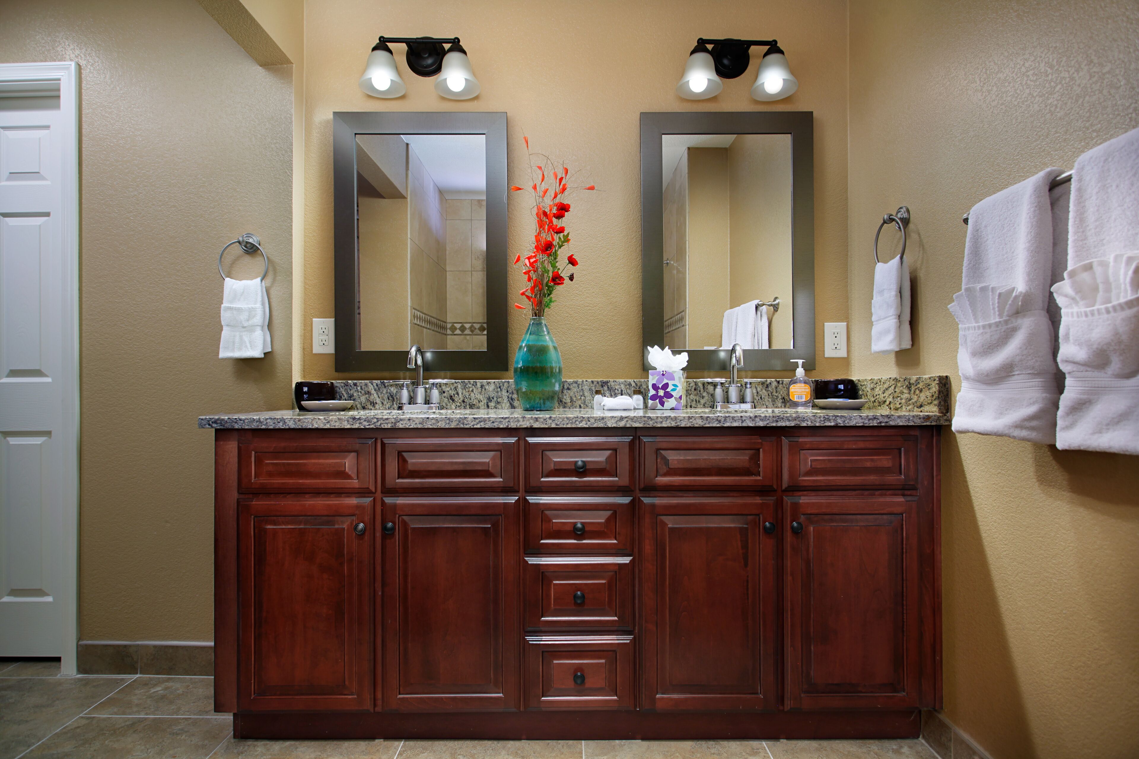 A bathroom with a double vanity, two mirrors, a vase with flowers, and towels on the wall.