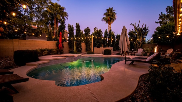 Piscina con jacuzzi, rodeada de sillas de descanso y sombrillas, luces colgantes y palmeras.