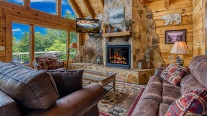 A log cabin interior with a stone fireplace, leather sofas, and a large window overlooking greenery. - City View Chalet, City View Chalet, Pool Table, Hot Tub, Free Attraction Tickets (Gatlinburg)