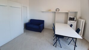 A room with a white ceiling fan, a blue sofa, a white table with blue chairs, a white radiator, and a shelf with a clock and a radio. - Vista Mare (Menton)