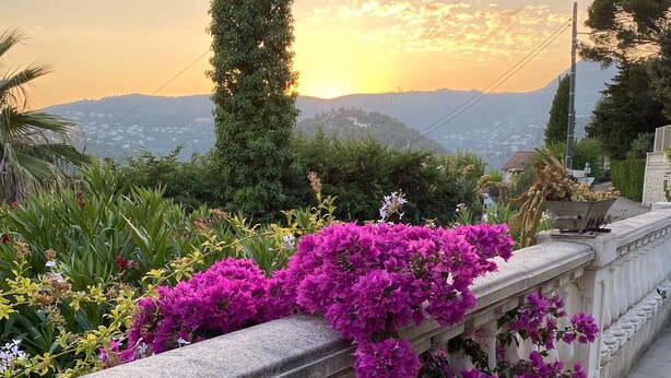 Ein Balkon mit leuchtend rosa Blumen und Blick auf einen Sonnenuntergang über einer Berglandschaft.