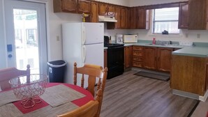 Une cuisine compacte avec des meubles en bois, un réfrigérateur blanc, un four à micro-ondes et une cuisinière. On y trouve une table à manger avec des chaises et une poubelle.
