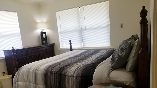 Une chambre à coucher comprenant un lit, une commode, une table de chevet et deux fenêtres équipées de stores.