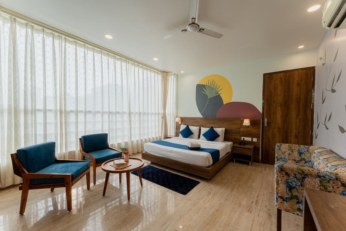 A hotel room with a bed, two blue armchairs, a small round table, a wooden floor, and a large window with sheer curtains.