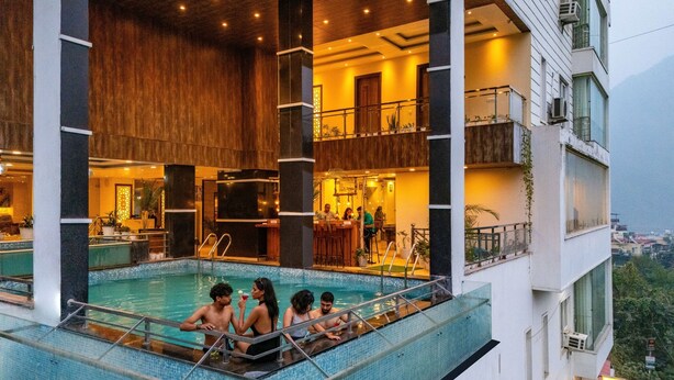 An indoor swimming pool with a view of the cityscape, surrounded by a modern building with multiple floors and balconies. - Moustache Rishikesh Luxuria - Hostel (Srinagar)