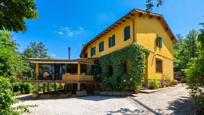 A yellow building with a balcony, surrounded by greenery and a gravel path.