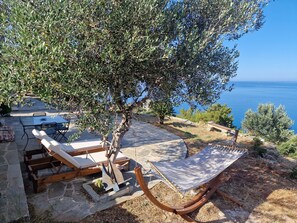 Un patio de piedra con una hamaca, sillones de descanso y una mesa dispuesto bajo un olivo con vistas al mar.