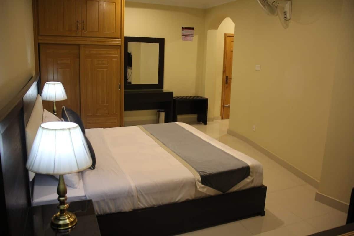 A hotel room with a bed, a nightstand with a lamp, a dresser, a mirror, and a door leading to another room.