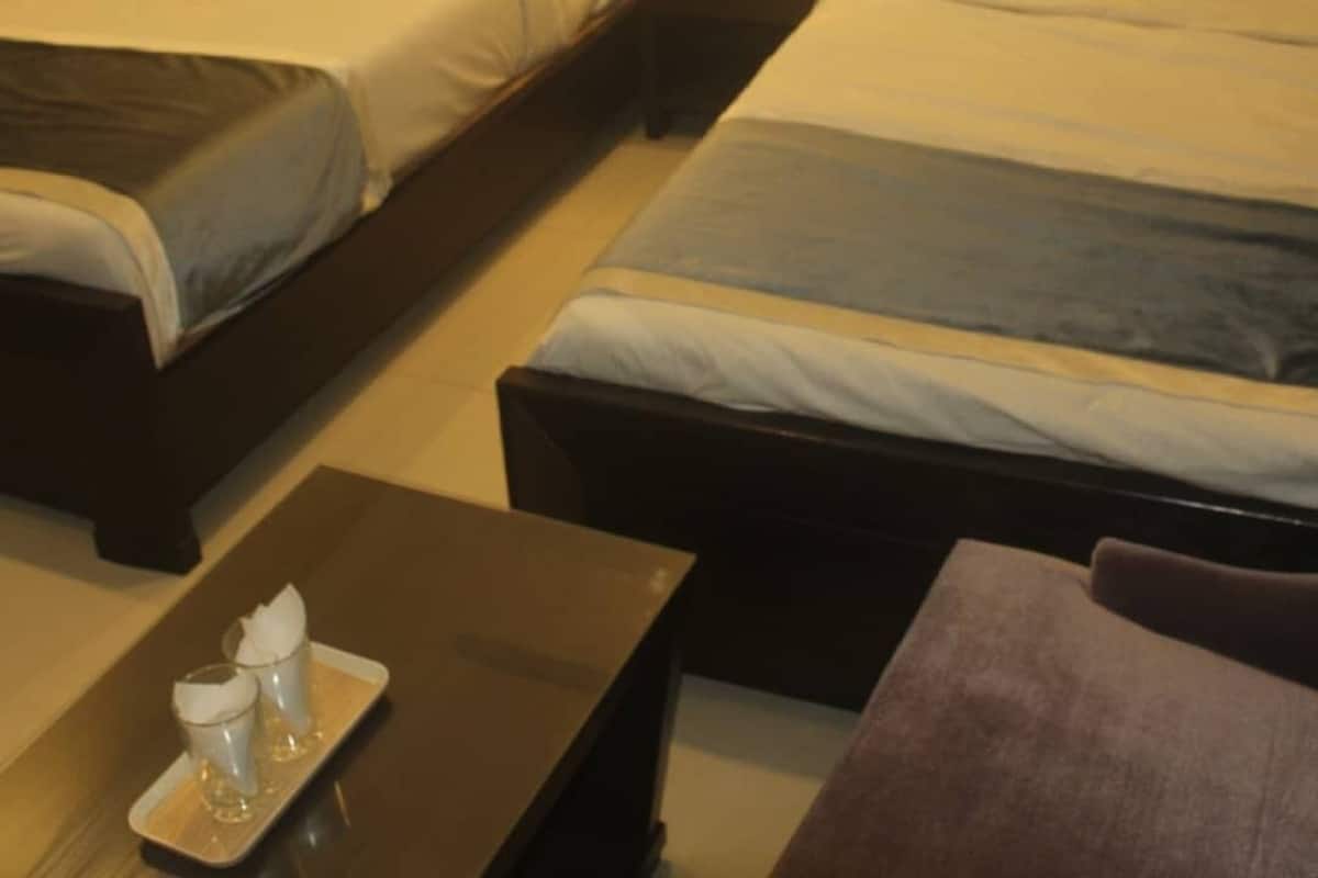 A hotel room with two single beds, a nightstand with a lamp, and a small table with a tray of glasses and a bottle.