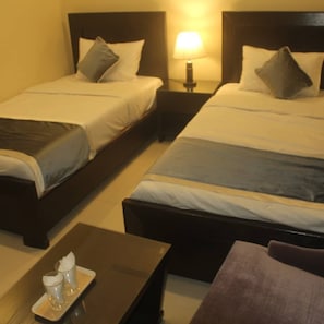 A hotel room with two single beds, a nightstand with a lamp, and a small table with a tray of glasses and a bottle. - Marjan Hotel (Rawalakot)