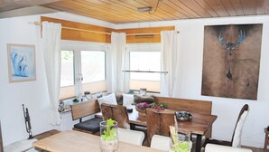 A dining area with a wooden table, white chairs, and a bench. There are two glass vases with green plants on the table.