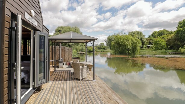 Een houten terras met een overdekte zithoek met uitzicht op een rustig meer, een houten gebouw met een geopende deur, en een bordje met de tekst 'Oak Lodge'.