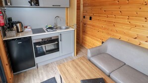Een gezellige kitchenette met een spoelbak, fornuis en kleine koelkast. In de woonkamer staat een grijze bank en een houten tafel.