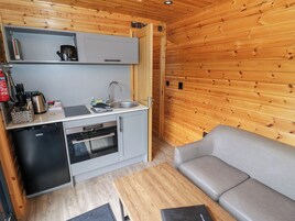 Cocina compacta con lavamanos, estufa y refrigerador pequeño. Hay un sofá gris y una mesa de madera en la sala.