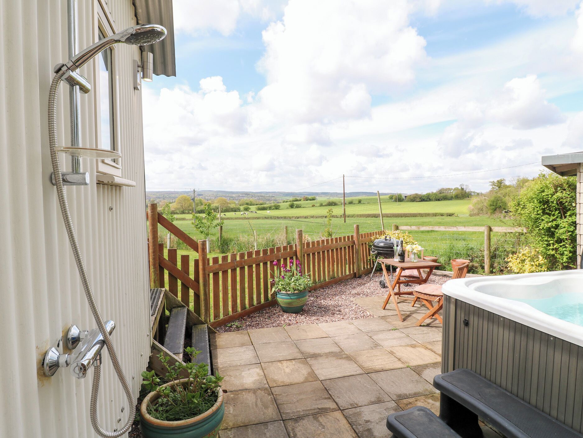 A patio with a hot tub, wooden fence, and a view of a green field.