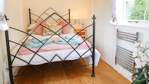 A bed with a diamond-patterned metal frame, a bedside table with a lamp, and a window with a view of greenery. - Shepherds Hut (Rugeley)