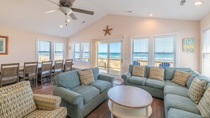 A spacious living room with a ceiling fan, large windows with a view of the ocean, and a dining area with wooden chairs and a table. - Carolina Sunshine - Nags Head MP 16, Oceanfront, Pool, Hot Tub, Pets OK, Flex Stays (Nags Head)