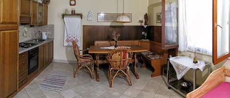 A compact kitchen with wooden cabinets, a dining table with chairs, a small window with curtains, and a bed with a pink bedspread.
