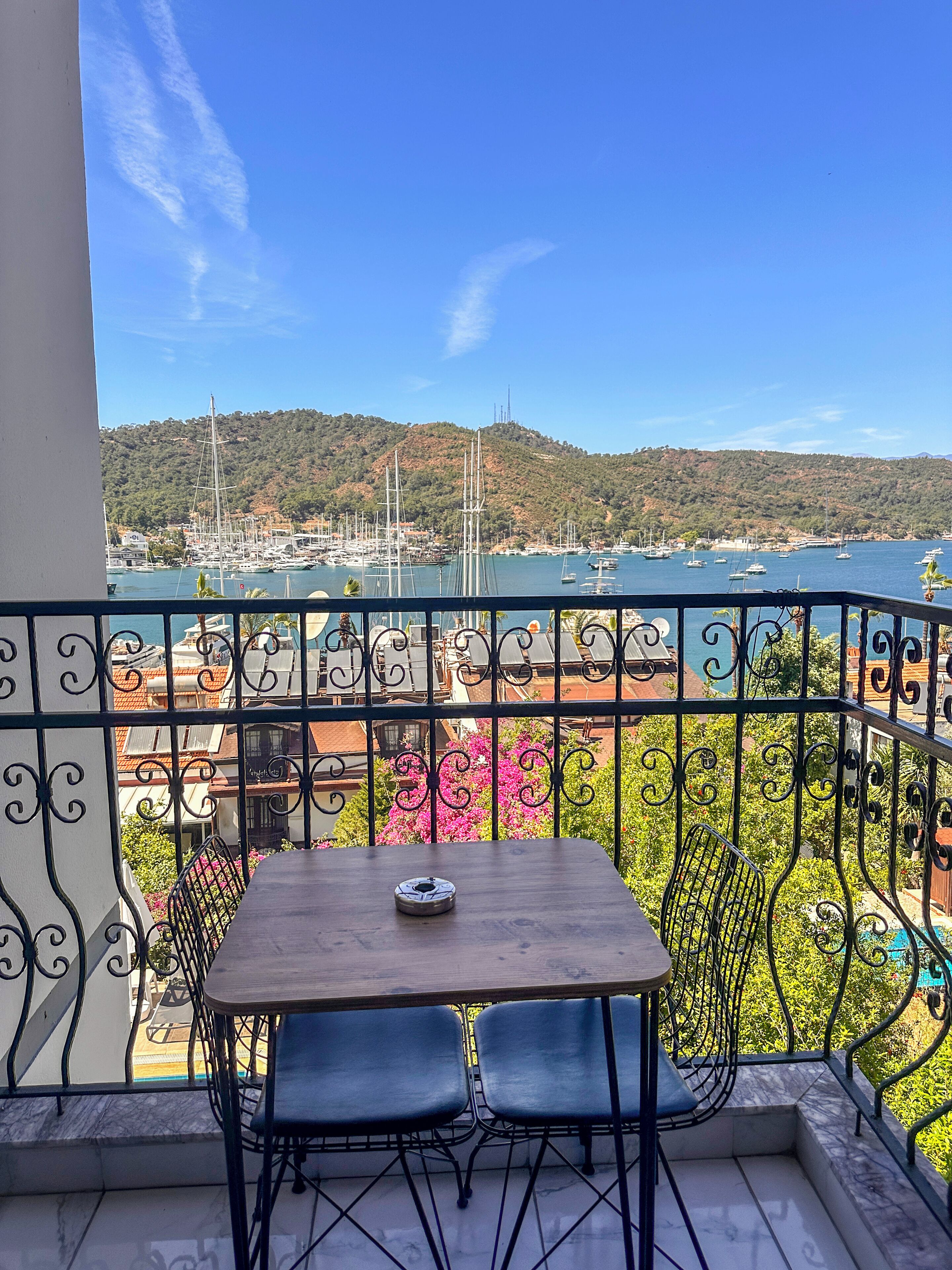 Un balcón con una mesa y dos sillas, con vistas a un puerto deportivo con barcos y un paisaje montañoso.