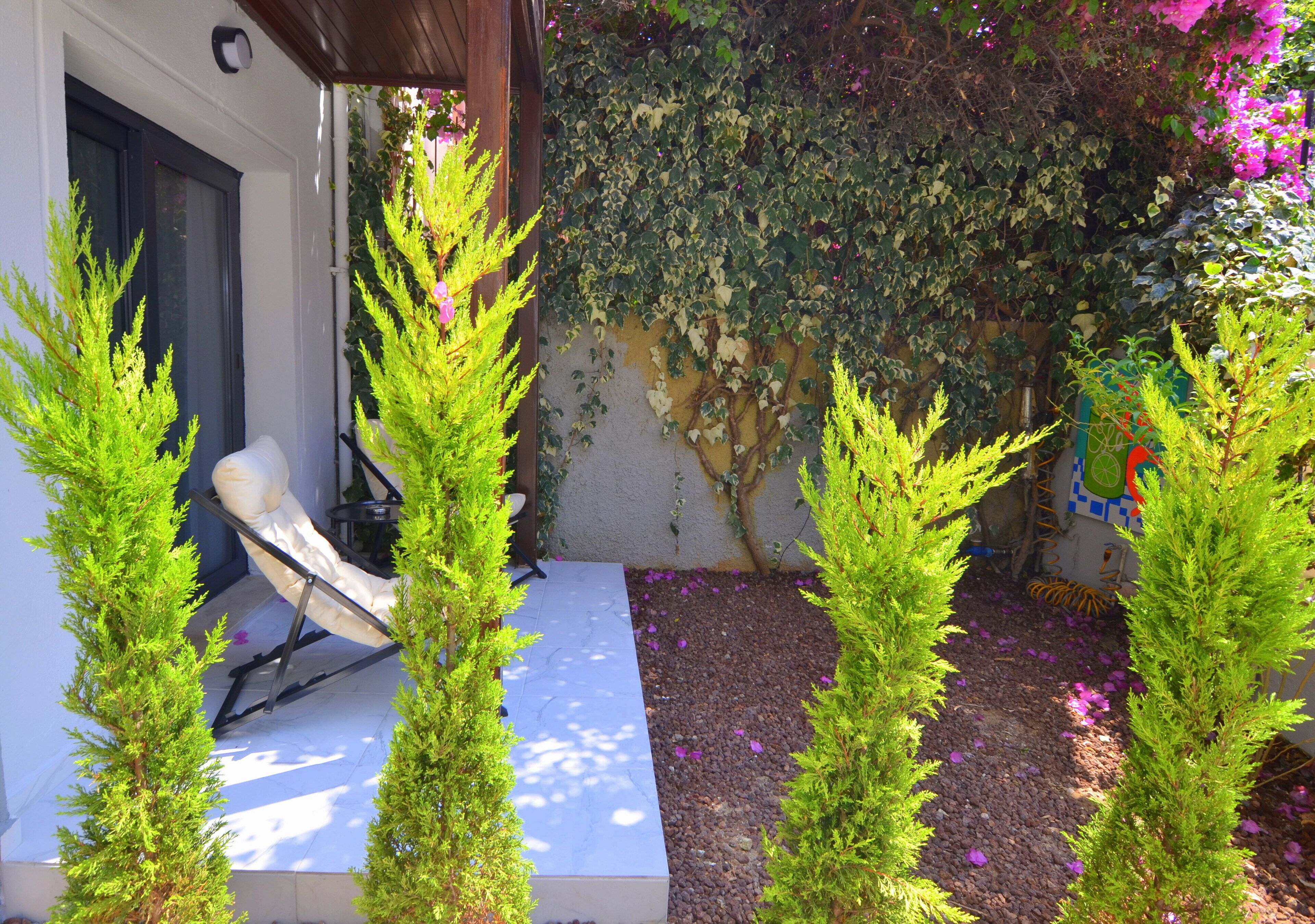 A covered outdoor seating area with a white bench, surrounded by green coniferous trees and a wall covered in climbing plants.