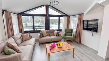Ein modernes Wohnzimmer mit Sofa, Sessel und Couchtisch, großen Fenstern mit Blick auf Boote und Hügel sowie einem Flachbildfernseher an der Wand.