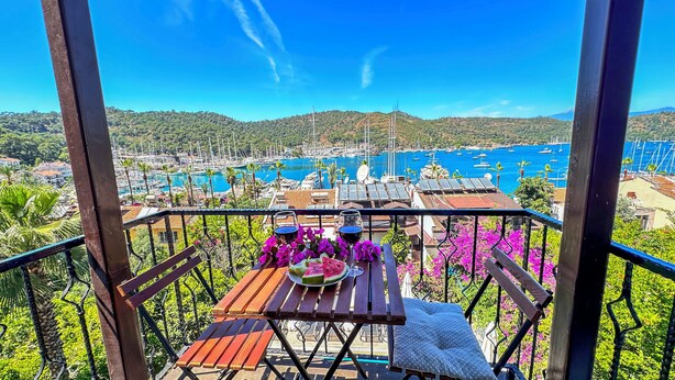 Ein Balkon mit einem Holztisch, zwei Gläsern Rotwein und einem Wassermelonenschnitz. Der Blick umfasst einen Yachthafen mit Booten und eine Berglandschaft.