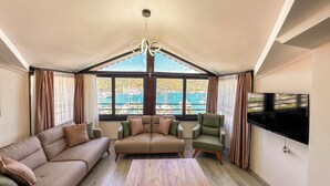 Ein geräumiges Wohnzimmer mit hoher Decke, einem Kronleuchter, einem großen Fernseher und Blick auf einen Yachthafen durch große Fenster.