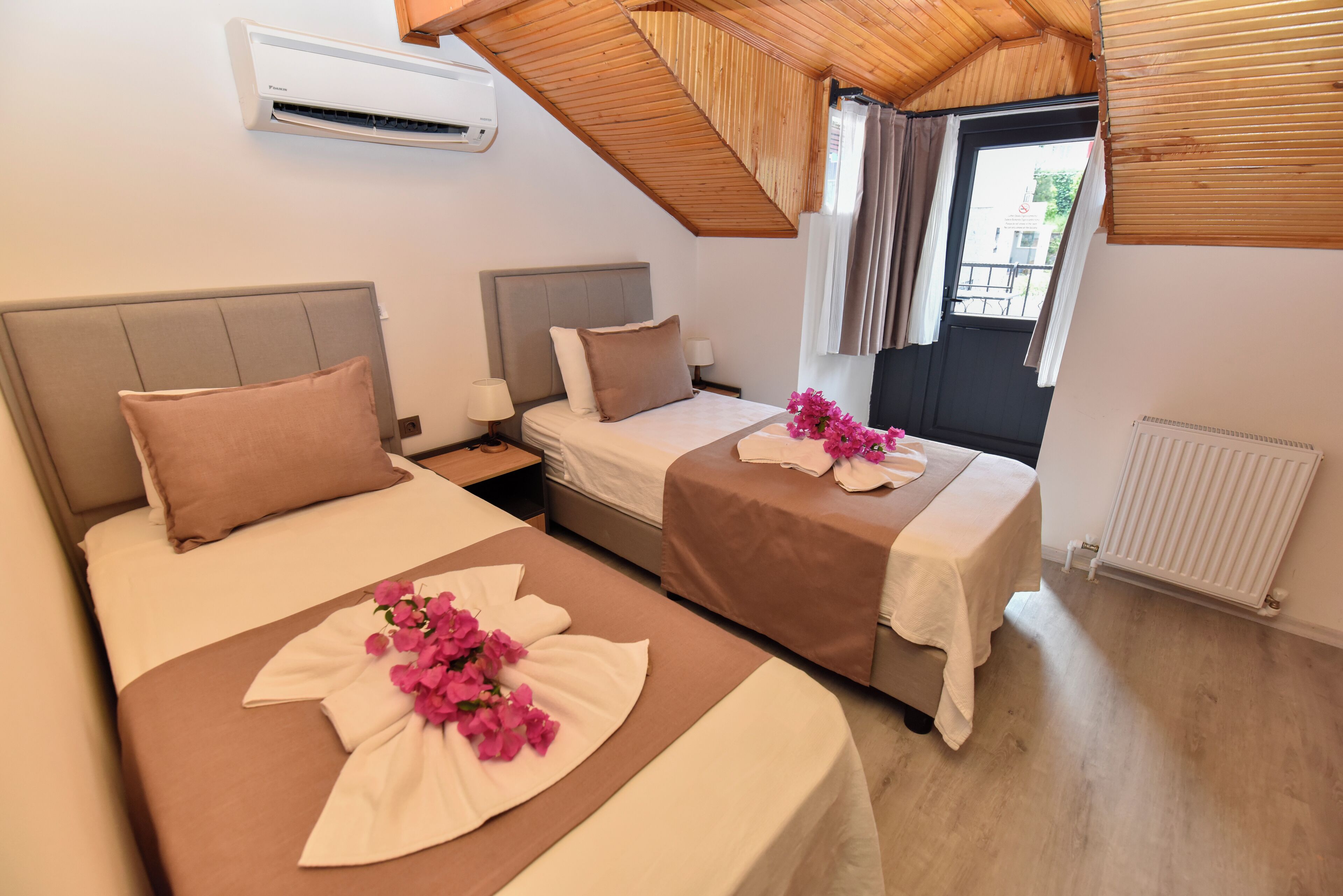 A hotel room with two beds, each with a pillow and a bedside table. There is an air conditioning unit on the wall, a window with curtains, and a radiator on the floor.