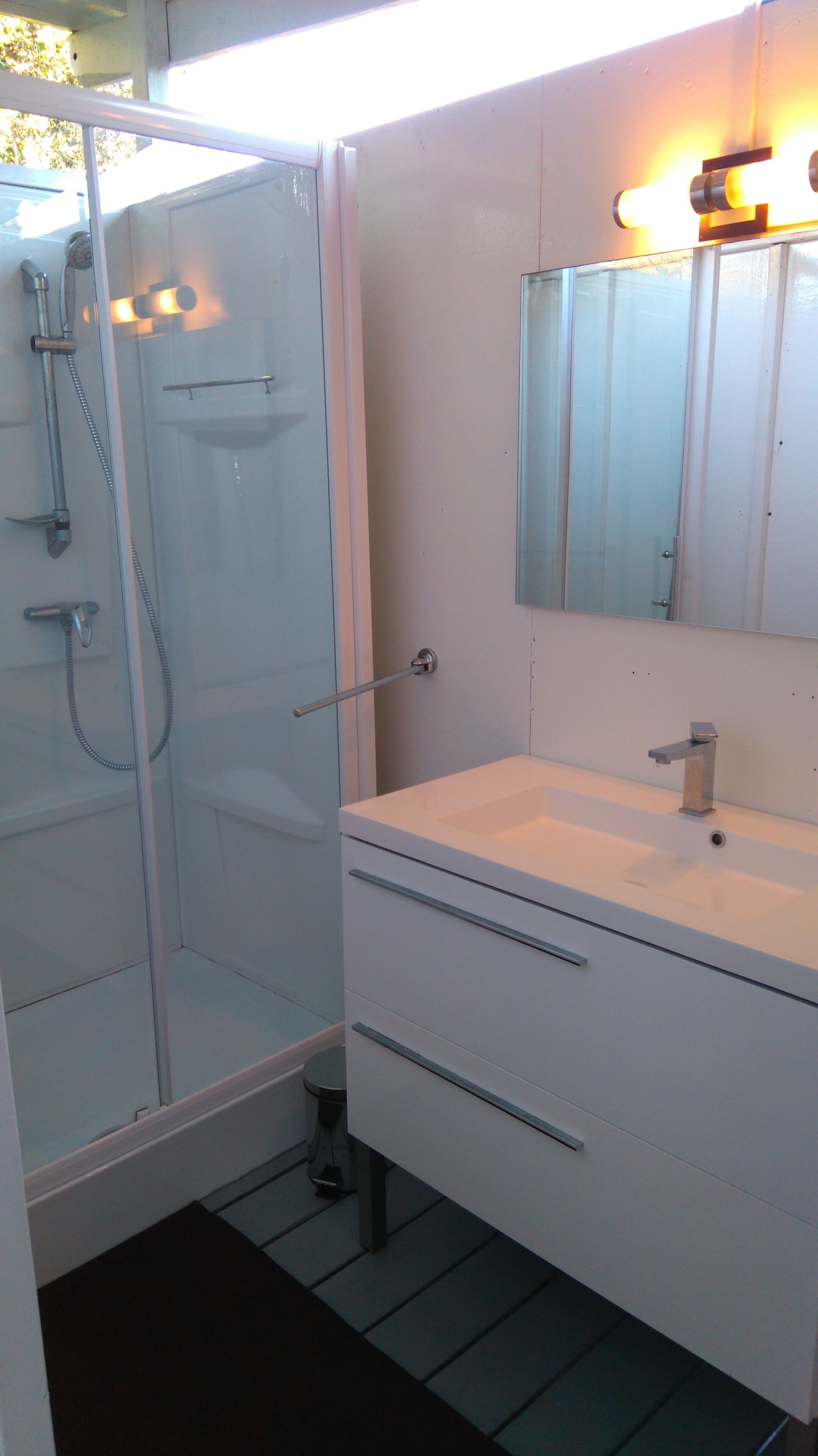 A modern bathroom with a glass shower enclosure, a white sink with a mirror above it, and a small black trash bin.