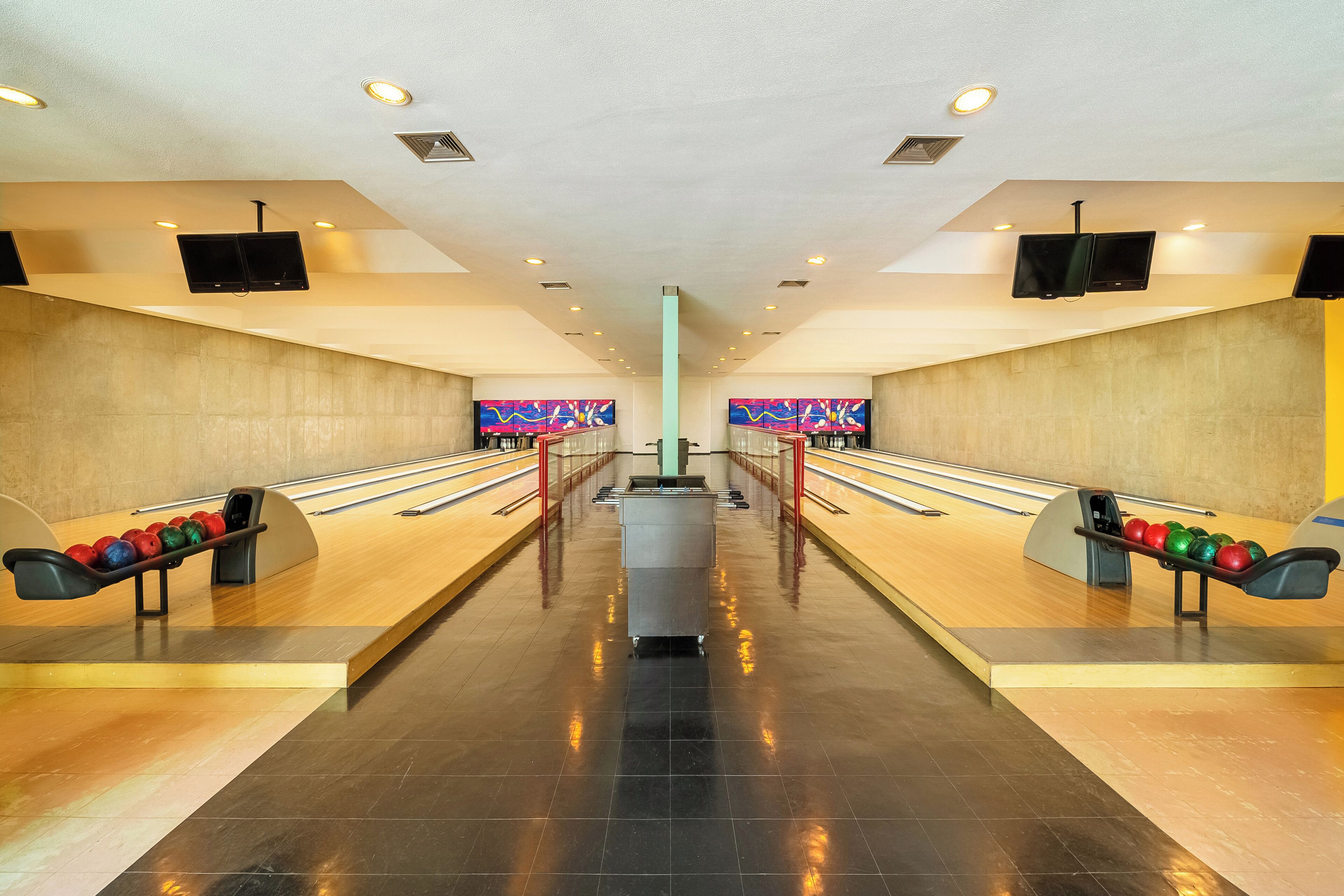 A bowling alley with two lanes, a central trash bin, and two televisions mounted on the walls.