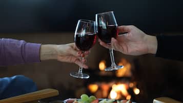 Two people toasting with wine glasses, a plate of cheese and grapes, and a fireplace in the background.
