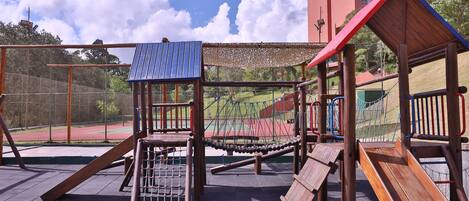 A playground with various play structures, including a slide and climbing areas, set against a backdrop of a sports field and buildings.