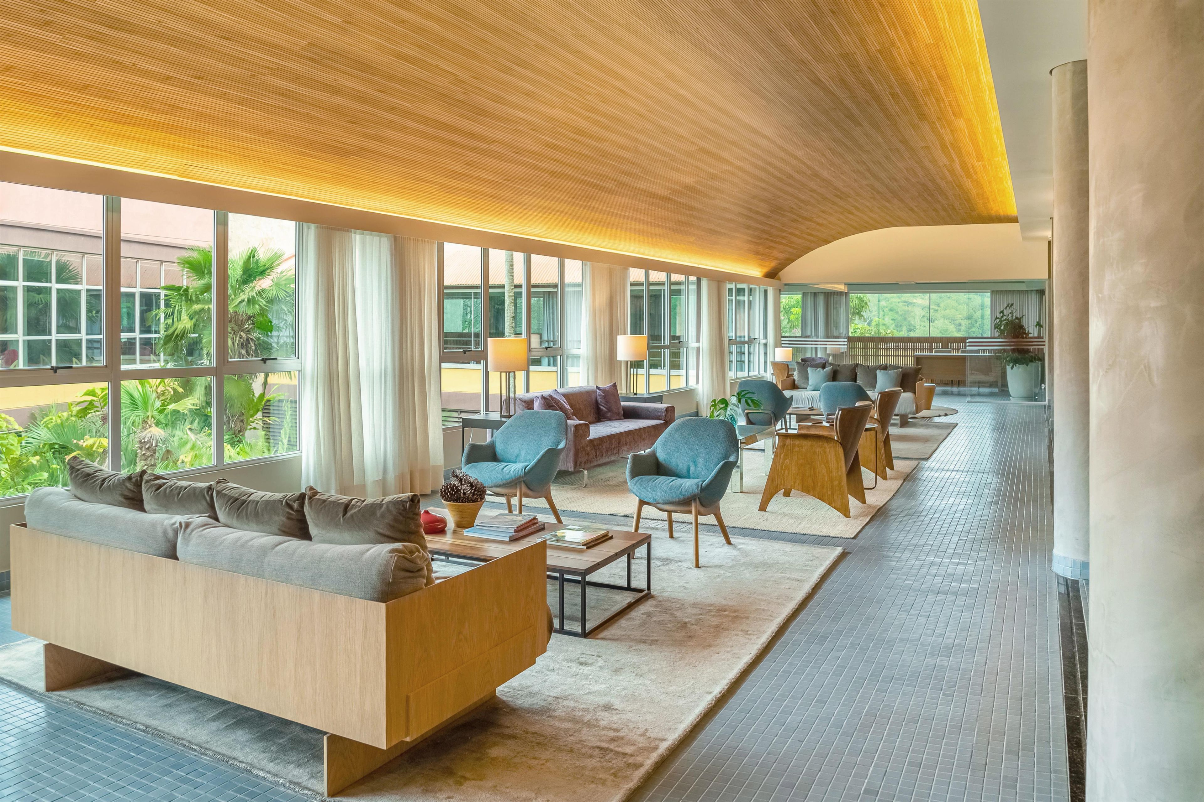 A spacious hotel lobby with a modern design, featuring a large seating area with sofas and armchairs, a coffee table, and a view of the outdoors through large windows.