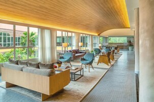 Um amplo hall de hotel com design moderno, com uma grande área de estar com sofás e poltronas, uma mesa de centro e vista para o exterior por grandes janelas.
