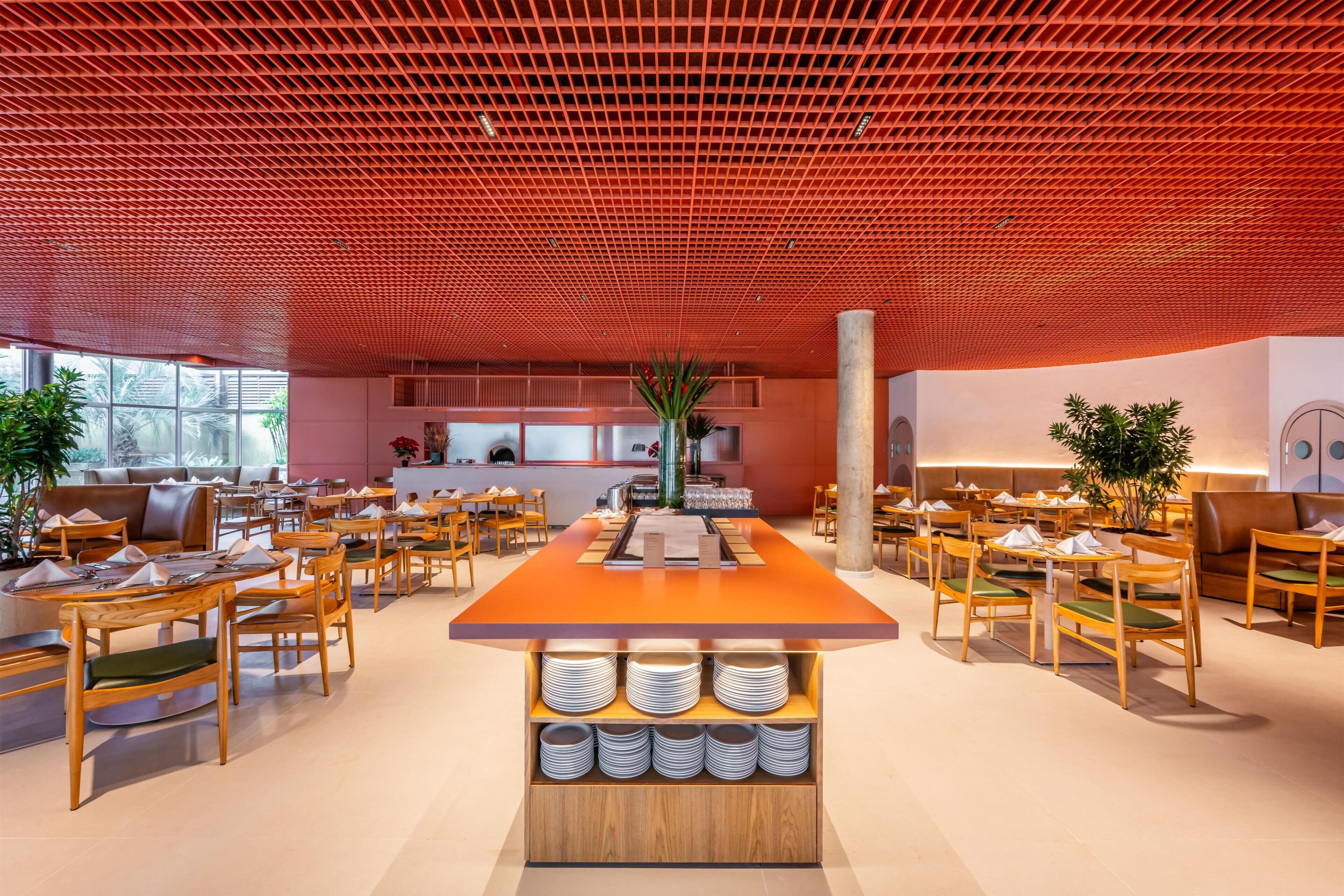 Een moderne eetruimte met een centraal eiland, houten tafels en stoelen, en een rood plafondrooster.