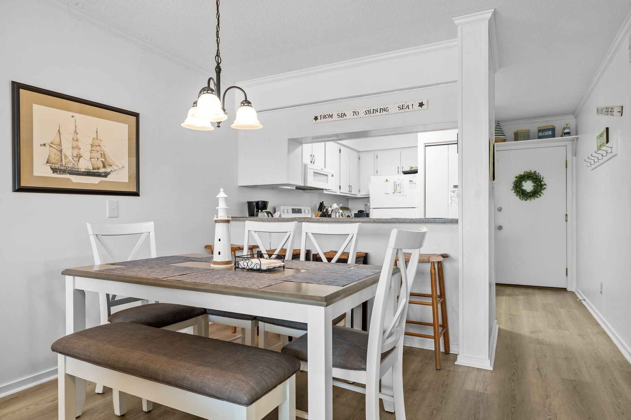 Un comedor con mesa de madera y sillas blancas, una cocina con armarios blancos y un cuadro en la pared.