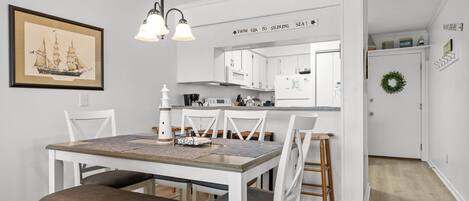 Un comedor con mesa de madera y sillas blancas, una cocina con armarios blancos y un cuadro en la pared.