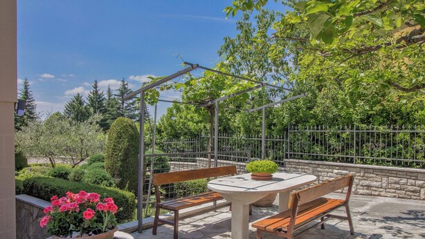 Eine Terrasse im Freien mit einem Steintisch und hölzernen Bänken, umgeben von einem Metallzaun und viel Grün.
