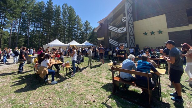 An outdoor event with people seated at tables under tents, a building with a modern design in the background, and trees surrounding the area. - Olimpo Sila Hotel (Taverna)