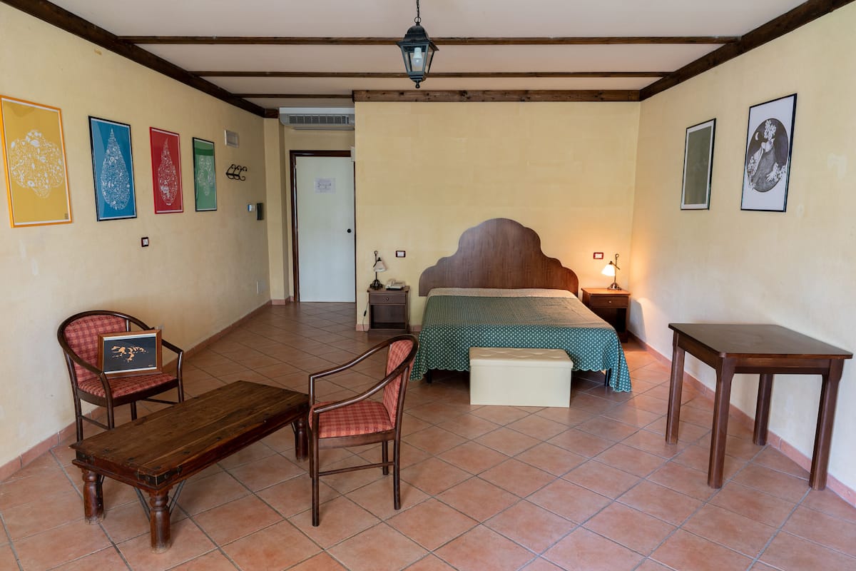 A room with a bed, two chairs, a table, and artwork on the walls.