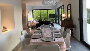 A modern dining area with a long table set for six, surrounded by white chairs. There is a chandelier above the table, and a sofa with cushions in the background. - Villa Exceptionnelle , Avec vue Panoramique (Venasque)