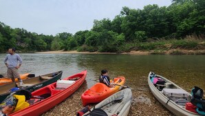 A row of kayaks and a paddleboard on a pebbly shore by a river, with a person standing nearby. - Kings River Bell Tent Glamping  (Eureka Springs)