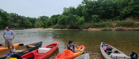 A row of kayaks and a paddleboard on a pebbly shore by a river, with a person standing nearby.