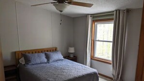 Une chambre à coucher comprenant un lit, un ventilateur de plafond, une table de chevet avec une lampe, une fenêtre avec des rideaux et une porte.