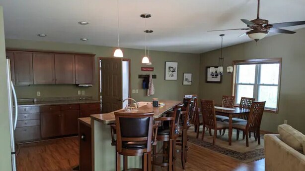 Une cuisine spacieuse avec des meubles en bois, un îlot central doté de tabourets, un coin repas avec une table et des chaises, et un ventilateur de plafond.