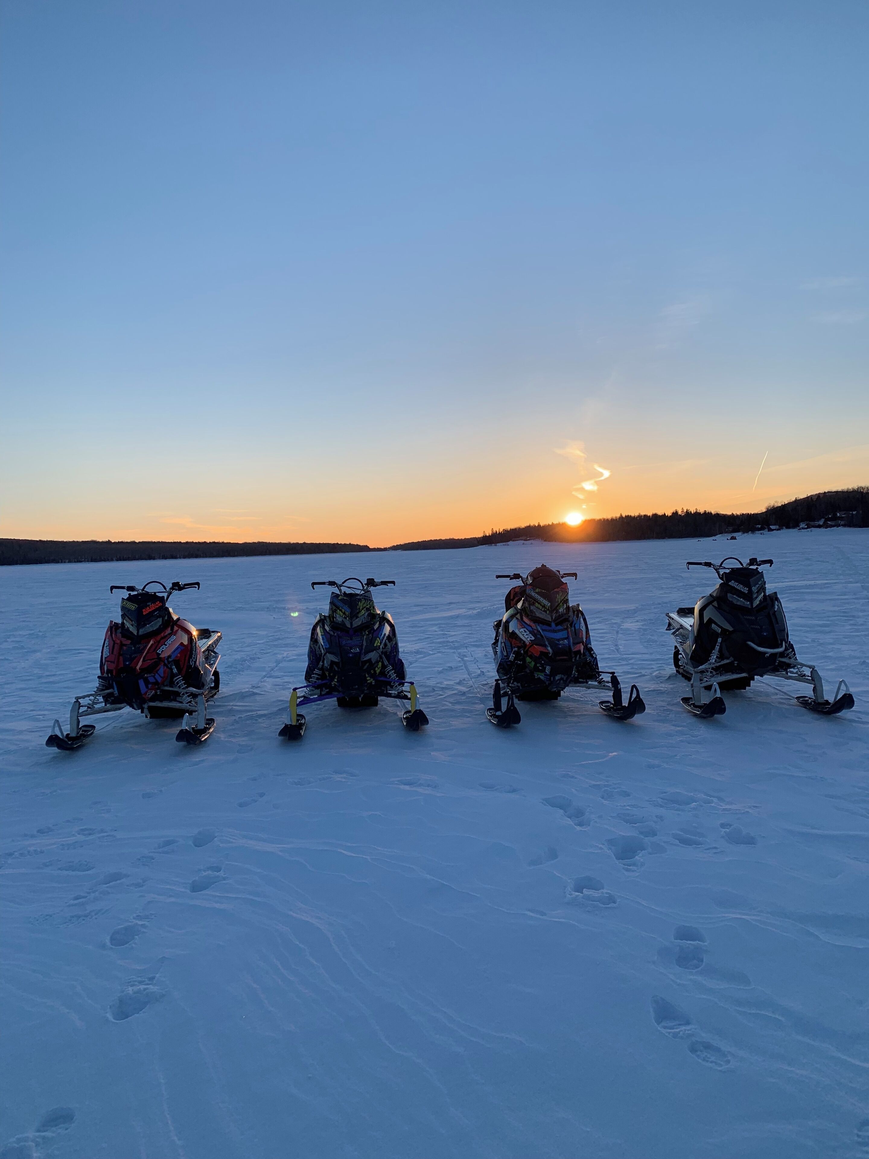 Quatre motoneiges sur un lac gelé, avec le soleil couchant en arrière-plan.